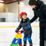 Un jeune enfant avec un casque et des protections patine sur une glace avec l'aide d'un adulte bienveillant.