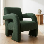 Fauteuil moderne au design sculptural recouvert d'un tissu bouclé vert forêt, posé sur un parquet en bois clair.